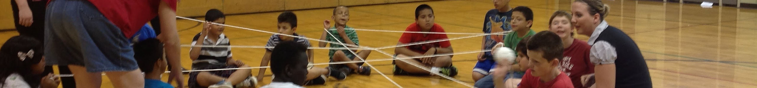 15 elementary children sit cross legged in a large circle on a wooden gymnasium floor. They are all wearing shorts and short sleeve shirts. One youth is holding a white ball. There is string being held by all students connecting them and making a spider web type pattern in the middle of the circle.