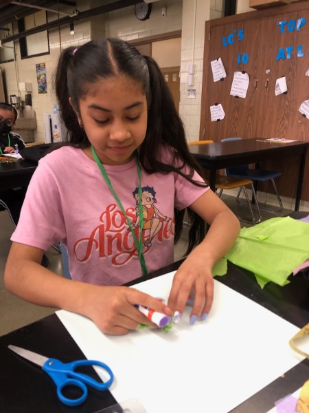 Young female sitting at a desk in a classroom. She is wearing a pink shirt that says Los Angeles with the character Betty Boop on it. The student has with long black hair in pony tails and is looking down at a paper. She is using a glue stick and gluing tissue paper to her plain white paper