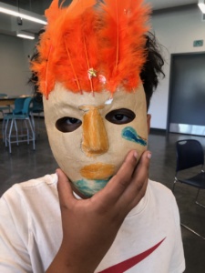 Young student holding a paper mache mask up to their face. The mask has yellow and blue markings with bright orange feathers sticking out at the top