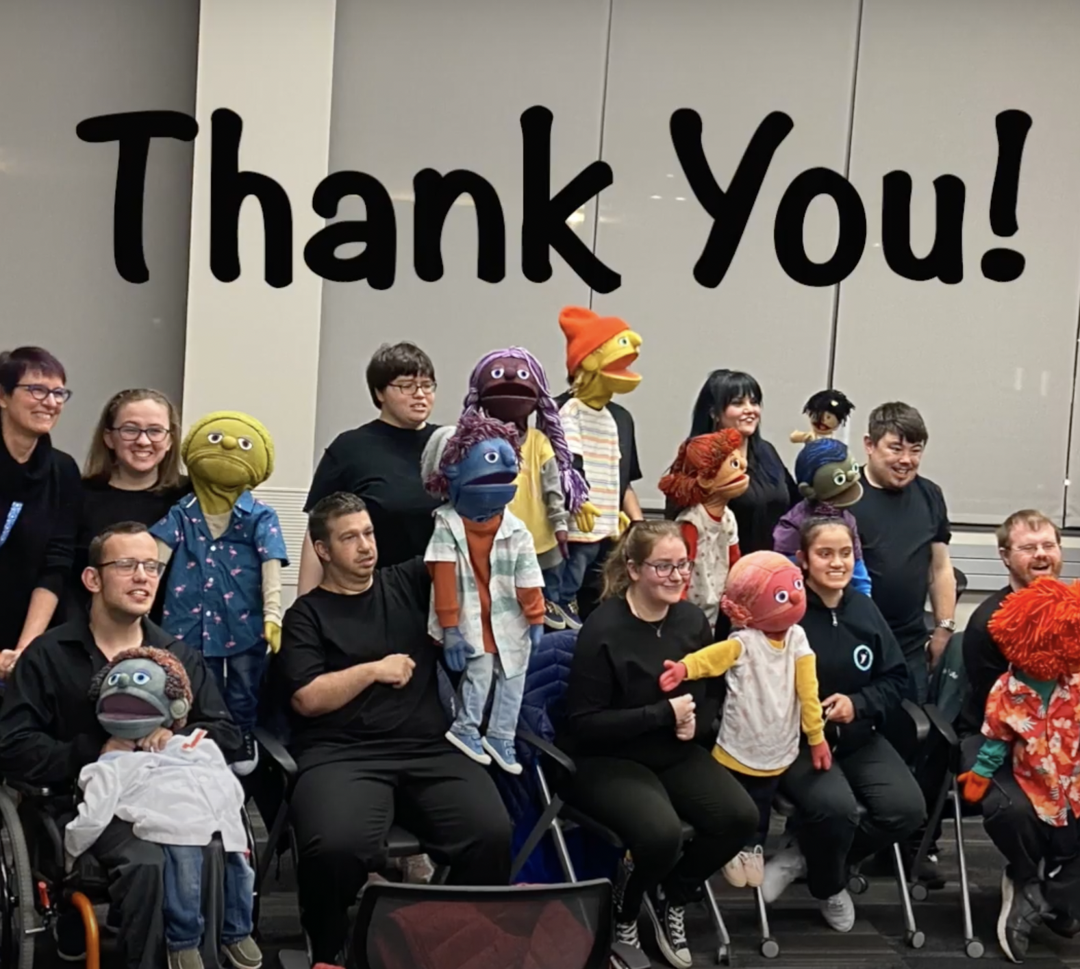 Approximately ten humans wearing black and each holding up a puppet. The puppets appear to be young and are all various bright colors. Text: Thank You